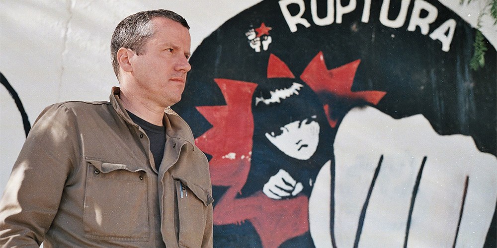 Presenter Carne Ross standing in front of an anarchist mural