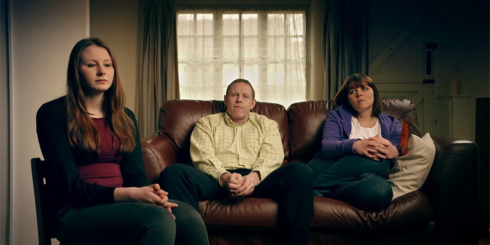 Daughter sits in chair in foreground while parents sit on sofa in the background