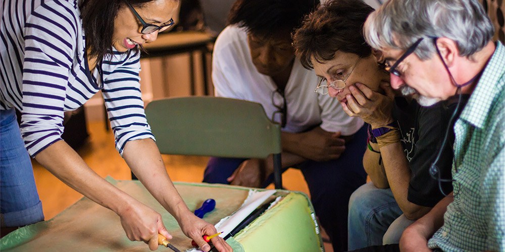 Female artist demonstrates how to upholster a chair to three onlookers
